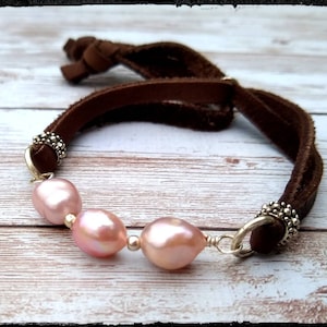 May include: A brown leather bracelet with three pink pearls and silver accents.