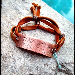May include: A brown leather bracelet with a copper plate that reads "Fearfully & Wonderfully Made". The bracelet has a small turquoise arrow charm.