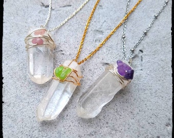 May include: Three crystal point necklaces with different colored stones wrapped in wire. The necklaces are on a gray textured surface. The stones are pink, green, and purple.