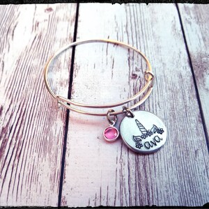 May include: Silver bangle bracelet with a charm featuring a unicorn silhouette and the name "Ava" engraved on it. The charm also has a pink rhinestone.
