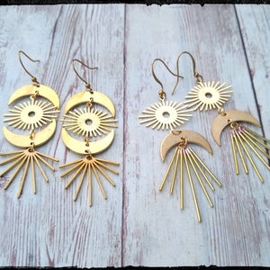 May include: Two pairs of gold tone earrings with a sun and moon design. The earrings feature a crescent moon with a sunburst design inside. The bottom of the earrings have a sunburst design with long rays.