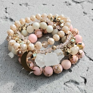 May include: A stack of beaded bracelets featuring a mix of pale pink, cream, and white beads with gold-toned accents. The bracelets include smooth round beads, textured marbled beads, clear faceted crystals, and a gold-toned crescent moon charm.