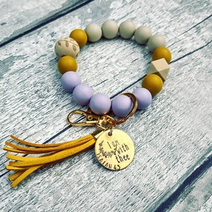 May include: A purple, beige, and mustard yellow beaded keychain with a gold tassel and a round gold charm that says "I am with thee Isaiah 43".