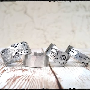 May include: Four silver metal rings with different engraved designs. The rings are on a white wooden surface.