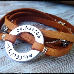May include: A brown leather wrap bracelet with a silver metal circle charm engraved with the coordinates "30.164930N -81.735310W".