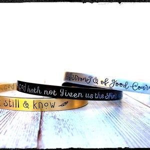 May include: Three metal bracelets with inspirational quotes engraved on them. The top bracelet is silver and says "Be Strong & of Good Courage". The middle bracelet is black and says "God hath not given us the Spirit". The bottom bracelet is gold and says "Be still & know".