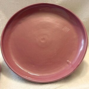 May include: A large, round, dusty rose-colored ceramic plate. The plate has a smooth, glossy finish and a slightly raised edge. The plate is part of a dinnerware set.