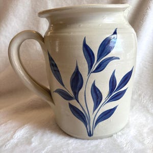 Peut inclure: Pichet en céramique de couleur crème avec une anse incurvée et un bec verseur. Le pichet est décoré d'un motif de feuilles bleues. Le pichet est probablement utilisé pour servir des boissons et a une esthétique rustique et artisanale.