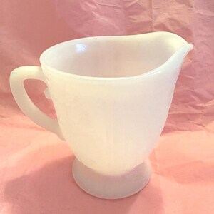 May include: A white, vintage-style creamer pitcher with a curved handle and a spout. The pitcher has a rounded base and a slightly textured surface. The creamer is made of opaque glass and is likely from the mid-20th century.
