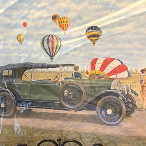 May include: Watercolor painting of a vintage green car with a black top, parked on a dirt road. Several colorful hot air balloons float in the sky above. A family stands near the car, observing the balloons.