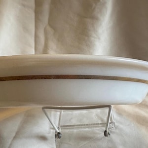 May include: A white, oval-shaped serving dish with a gold band around the middle. The dish is resting on a white metal stand, and the background is a light beige fabric.