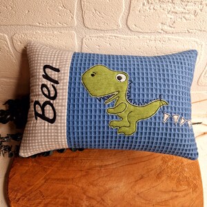 Könnte beinhalten: Ein blaues und graues Waffel-Kissen mit einem grünen Dinosaurier-Applique und dem Namen "Ben" auf der Seite bestickt.