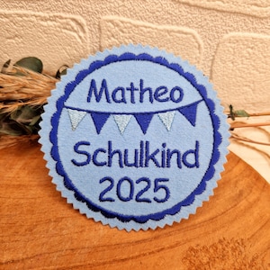 Könnte beinhalten: Blauer und weißer Filz-Kreis mit dem Text "Matheo Schulkind 2025" und einer Girlande aus blauen und weißen Flaggen.