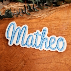Könnte beinhalten: Weißer Filz-Patch mit dem Namen "Matheo", der in hellblauem Garn gestickt ist.