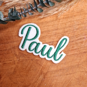 Könnte beinhalten: Weißer Filznamens-Patch mit dem Namen "Paul", der in grünem Garn gestickt ist.