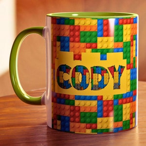 May include: A white ceramic mug with a colourful brick pattern and the name "CODY" in bold letters. The mug has a green handle and interior, sitting on a wooden surface. The design features red, blue, yellow, and green blocks.