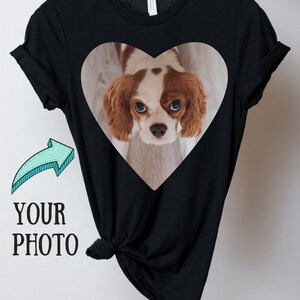 May include: A black short-sleeved t-shirt featuring a heart-shaped graphic containing a photograph of a brown and white Cavalier King Charles Spaniel. To the left of the heart graphic, there is a light blue arrow pointing toward the text YOUR PHOTO.