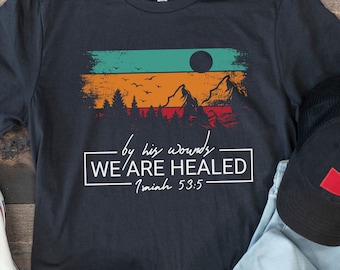 May include: Dark gray t-shirt with a graphic of a mountain scene in orange, green, and red tones. The text "by his wounds WE ARE HEALED Isaiah 53:5" is printed in white. Includes a pair of white sneakers, a black cap, and blue jeans.