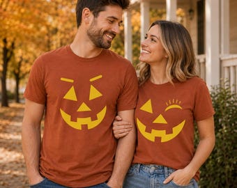May include: Two rust-colored short-sleeve t-shirts featuring yellow jack-o'-lantern face graphics. One shirt displays a standard smiling pumpkin face, while the other features a winking pumpkin face with a single triangular eye and eyelashes.
