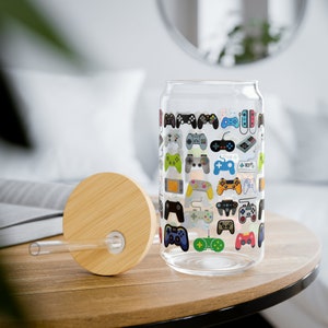 May include: A clear glass can-shaped tumbler with a bamboo lid and straw. The glass is decorated with colorful illustrations of various video game controllers. The tumbler sits on a wooden table.