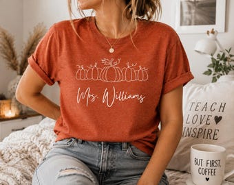 Könnte beinhalten: Ein rostfarbenes Kurzarm-T-Shirt mit einer weißen Strichgrafik von fünf nebeneinander angeordneten Kürbissen über dem in Schreibschrift verfassten Text "Mrs. Williams". Das T-Shirt hat einen Rundhalsausschnitt und wird mit einer blauen Jeans getragen.