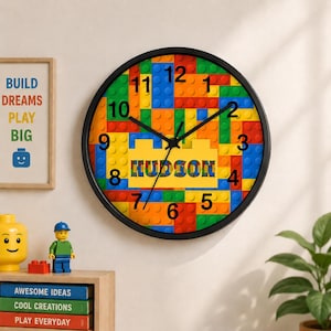 May include: A round wall clock with a black frame. The clock face is designed to resemble colorful toy building blocks, with the name "HUDSON" in a yellow block. Numbers are placed around the clock face.