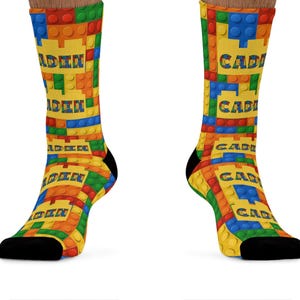 K&ouml;nnte beinhalten: Ein Paar Socken mit einem wiederkehrenden Muster aus ineinandergreifenden Kunststoff-Bausteinen in Rot, Blau, Gr&uuml;n und Gelb. Das Design enth&auml;lt den Namen CADEN, der in einer fetten, blockartigen Schrift auf gelben Feldern gedruckt ist.