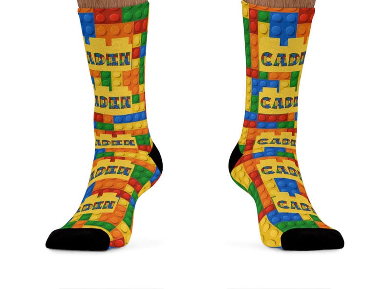 K&ouml;nnte beinhalten: Ein Paar Socken mit einem wiederkehrenden Muster aus ineinandergreifenden Kunststoff-Bausteinen in Rot, Blau, Gr&uuml;n und Gelb. Das Design enth&auml;lt den Namen CADEN, der in einer fetten, blockartigen Schrift auf gelben Feldern gedruckt ist.