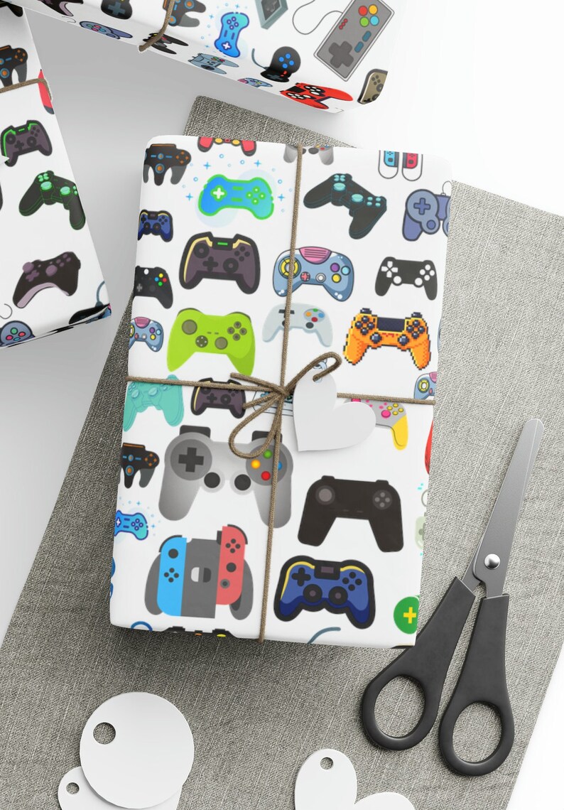 May include: White wrapping paper featuring a pattern of colourful video game controllers. The wrapped gift is tied with twine and a heart-shaped tag. Scissors and gift tags are also visible.