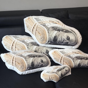 May include: A collection of five soft pillows shaped like stacks of one hundred dollar bills. Each pillow features a realistic print of Benjamin Franklin and is secured with a printed rubber band design, varying in size from large to small.