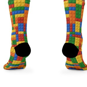 K&ouml;nnte beinhalten: Ein Paar Socken mit einem lebendigen, sich wiederholenden Muster aus ineinandergreifenden Kunststoff-Bausteinen in den Prim&auml;rfarben Rot, Blau, Gelb und Gr&uuml;n. Die Socken haben einfarbig schwarze Fersen und eine strukturierte Noppenoberfl&auml;che.