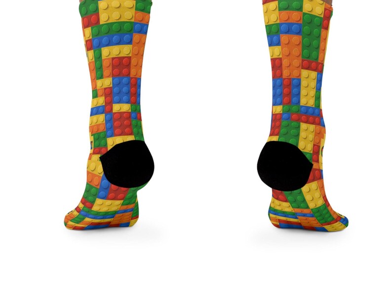 K&ouml;nnte beinhalten: Ein Paar Socken mit einem lebendigen, sich wiederholenden Muster aus ineinandergreifenden Kunststoff-Bausteinen in den Prim&auml;rfarben Rot, Blau, Gelb und Gr&uuml;n. Die Socken haben einfarbig schwarze Fersen und eine strukturierte Noppenoberfl&auml;che.