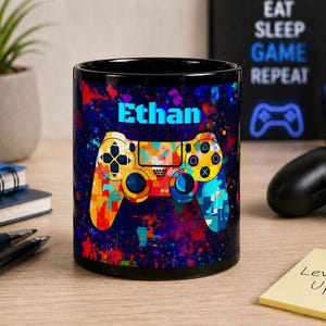 May include: Black coffee mug with a colorful controller design and the name "Ethan" in blue. The controller is a vibrant mix of yellow, orange, blue, and green. The mug has a dark blue and red splatter background.