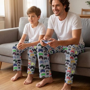 May include: Two people wearing matching gray pajama pants with a colorful video game controller pattern. They are sitting on a gray couch and wearing white t-shirts. The pajama pants feature a variety of controller designs in multiple colors.