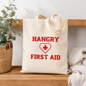 Peut inclure: Sac fourre-tout crème avec les mots "HANGRY" au-dessus d'un cœur rouge avec une croix, et "FIRST AID" en dessous. Le sac a un design simple et est fait d'un tissu robuste avec des poignées assorties.