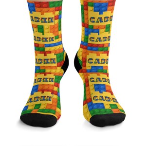 K&ouml;nnte beinhalten: Ein Paar Socken mit einem sich wiederholenden Muster aus bunten, ineinandergreifenden Kunststoff-Bausteinen in Rot, Blau, Gr&uuml;n und Gelb. Der Name CADEN ist in fetten blauen Buchstaben auf gelben rechteckigen Bl&ouml;cken im Design gedruckt.