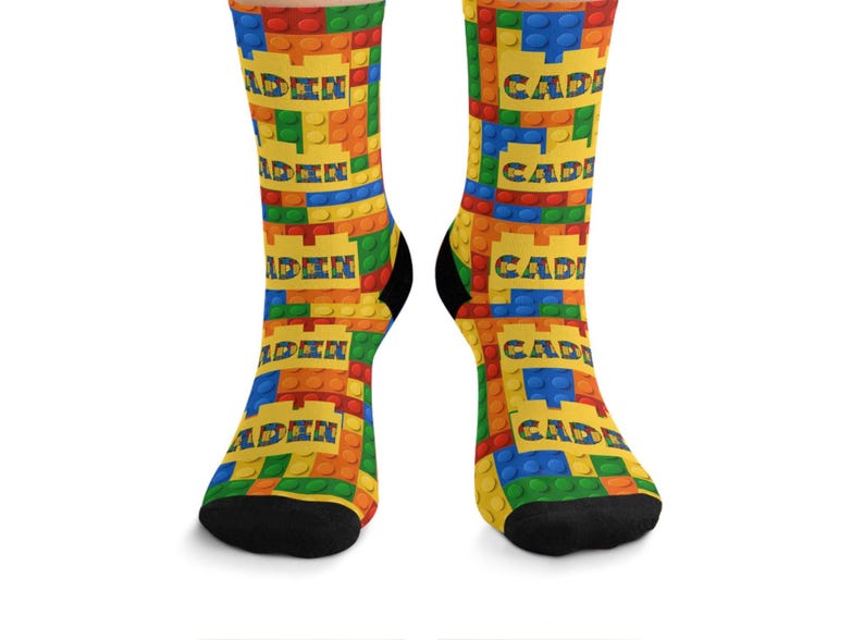 K&ouml;nnte beinhalten: Ein Paar Socken mit einem sich wiederholenden Muster aus bunten, ineinandergreifenden Kunststoff-Bausteinen in Rot, Blau, Gr&uuml;n und Gelb. Der Name CADEN ist in fetten blauen Buchstaben auf gelben rechteckigen Bl&ouml;cken im Design gedruckt.