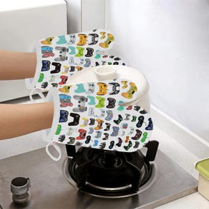 K&ouml;nnte beinhalten: Ein Paar wei&szlig;e Ofenhandschuhe mit einem bunten Muster aus Videospielcontrollern. Die Handschuhe sind aus hitzebest&auml;ndigem Material gefertigt und sch&uuml;tzen Ihre H&auml;nde beim Kochen oder Backen.