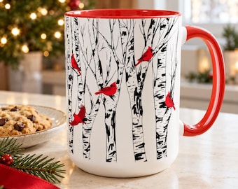 May include: A white ceramic mug featuring a black and white illustration of birch trees with several bright red cardinal birds perched on the branches. The mug has a glossy red interior and a matching red handle, standing on a light surface.