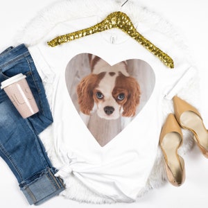 May include: A white t-shirt featuring a heart-shaped graphic of a brown and white Cavalier King Charles Spaniel, paired with blue denim jeans, a pink polka-dot travel mug, and beige pointed-toe heels on a white faux fur background.