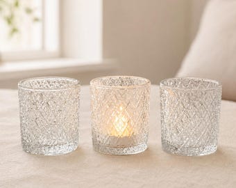 May include: Three clear glass votive candle holders with a textured diamond pattern. One holder contains a lit candle, casting a warm glow. The other two are empty. The candle holders are on a neutral-colored surface.
