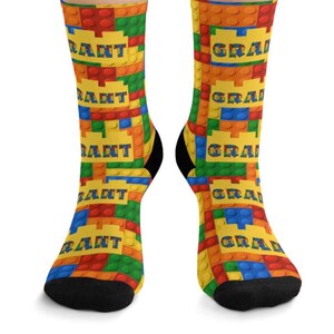 K&ouml;nnte beinhalten: Ein Paar Socken mit einem sich wiederholenden Muster aus bunten, ineinandergreifenden Kunststoff-Bausteinen in Rot, Blau, Gr&uuml;n und Orange. Das Design enth&auml;lt gelbe rechteckige Abschnitte mit dem Namen GRANT in blauen Buchstaben.