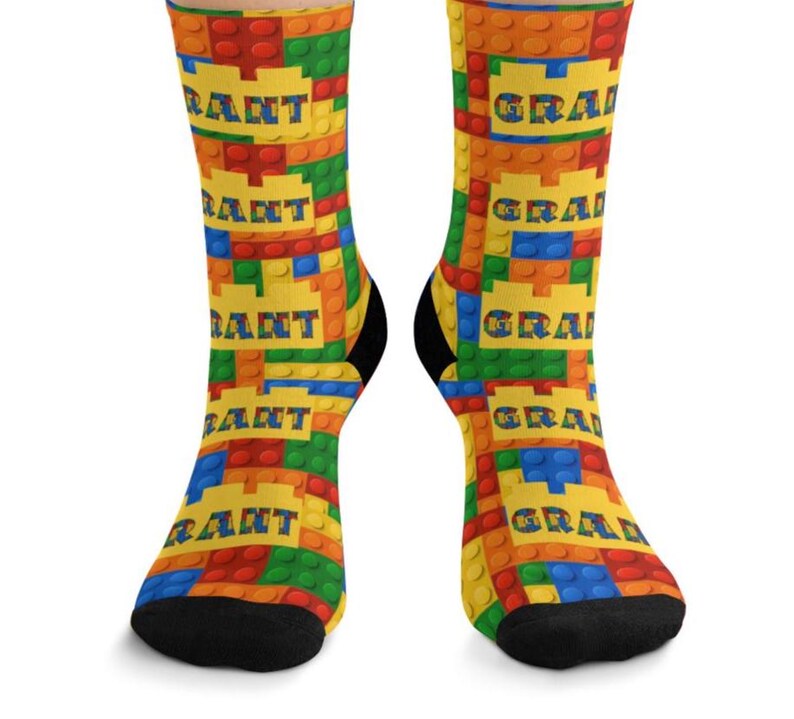 K&ouml;nnte beinhalten: Ein Paar Socken mit einem sich wiederholenden Muster aus bunten, ineinandergreifenden Kunststoff-Bausteinen in Rot, Blau, Gr&uuml;n und Orange. Das Design enth&auml;lt gelbe rechteckige Abschnitte mit dem Namen GRANT in blauen Buchstaben.