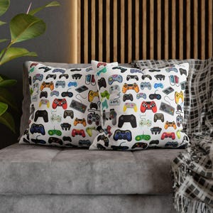 May include: Two square throw pillows with a white background featuring a repeating pattern of various colorful video game controllers and handheld consoles in shades of red, blue, yellow, green, black, and grey scattered across the fabric.