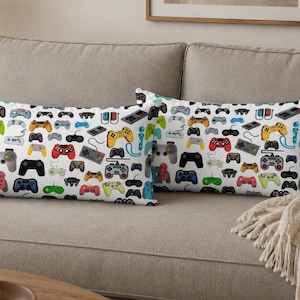 May include: Two rectangular throw pillows featuring a repeating pattern of various video game controllers in multiple colors including red, blue, green, yellow, black, and grey on a white background. The controllers vary in shape and style.