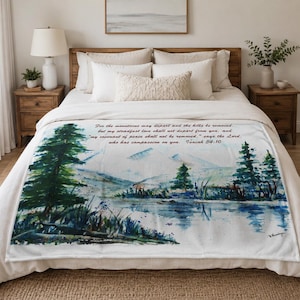 May include: A white fleece blanket featuring a watercolor landscape of a lake, pine trees, and mountains. The text reads: For the mountains may depart and the hills be removed, but my steadfast love shall not depart from you, Isaiah 54:10.