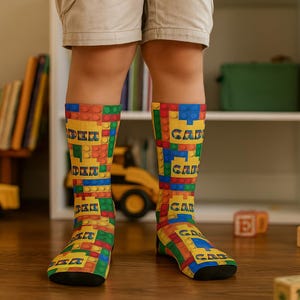 Peut inclure: Des chaussettes montantes avec un motif de briques colorées. Les chaussettes présentent les mots "DER" et "GAD" dans un style de lettres en blocs, avec un mélange de briques jaunes, rouges, vertes, bleues et oranges.