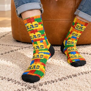 K&ouml;nnte beinhalten: Ein Paar Socken mit einem sich wiederholenden Muster aus ineinandergreifenden Kunststoff-Bausteinen in den Prim&auml;rfarben Rot, Gelb, Gr&uuml;n und Blau. Das Wort CHAD ist in einer fetten, blockartigen Schrift auf den gelben Bereichen gedruckt.