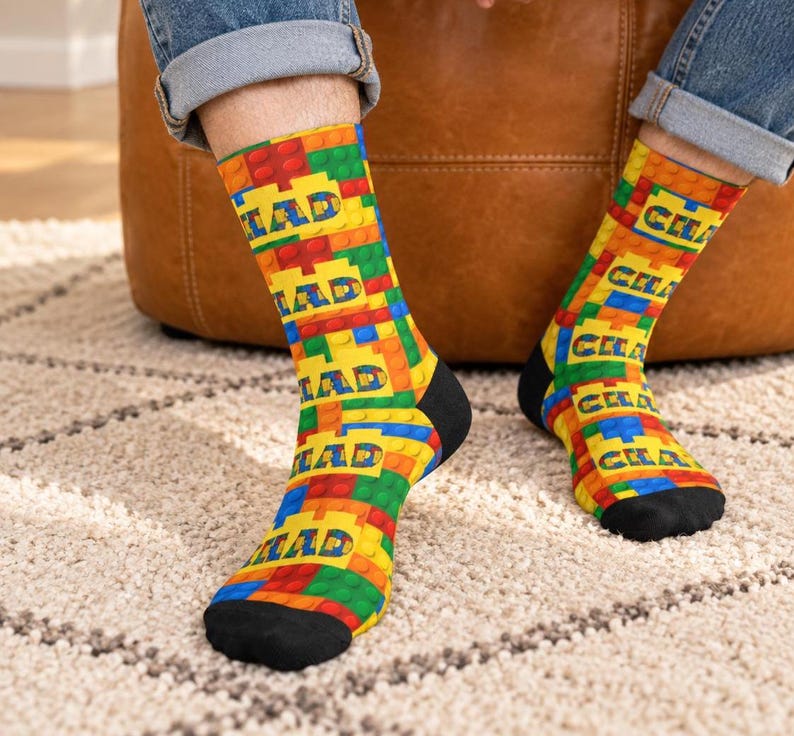 K&ouml;nnte beinhalten: Ein Paar Socken mit einem sich wiederholenden Muster aus ineinandergreifenden Kunststoff-Bausteinen in den Prim&auml;rfarben Rot, Gelb, Gr&uuml;n und Blau. Das Wort CHAD ist in einer fetten, blockartigen Schrift auf den gelben Bereichen gedruckt.