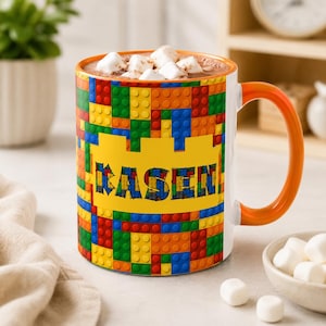May include: A ceramic mug with a vibrant, multi-colored pattern resembling interlocking plastic building blocks. The mug features a bright yellow central rectangular panel with the name KASEN printed in bold, blocky, multi-colored letters.